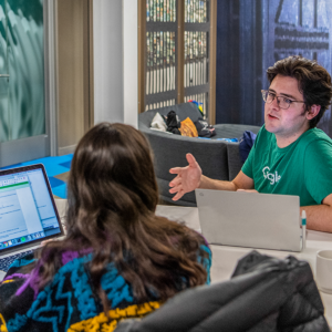 A man and a women, each with laptops open, discuss something in a professional setting.
