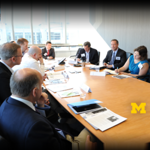 A boardroom with business people discussing around a conference table. The University of Michigan logo is in the lower corner.