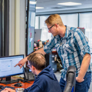A person overlooks another as he works on a computer in an office setting. The person overlooking is pointing at the screen.